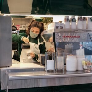 Masked barista making coffee in outdoor street café setting.